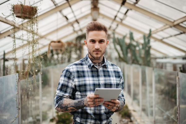 Businessman selling flowers and seedlings, standing in greenhouse, holding tablet. Small greenhouse business.