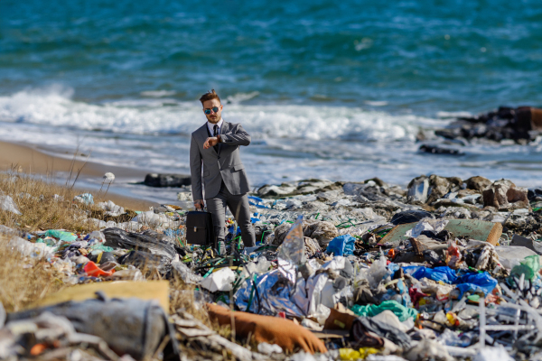 Fashionable modern businessman looking at watch, checking time standing on pile of waste on beach. Consumerism versus pollution concept. Corporate social responsiblity in business.