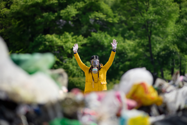 Female activist with gas mask holding hands up, standing on landfill, large pile of waste, environmental concept and eco activism.