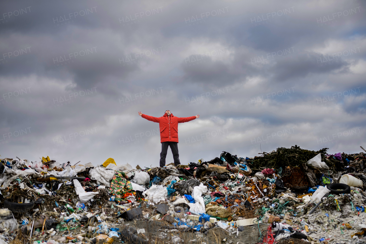 Activist in red jacket standing on landfill, on large pile of waste with dark clouds above. Environmental concept and eco activism.