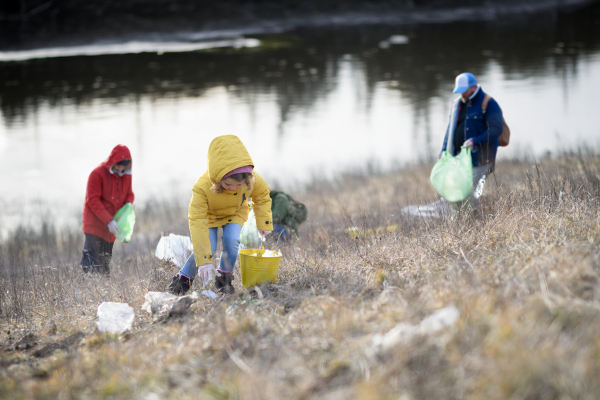 Young girl among eco activists picking up litter along riverbank, in nature, environmental pollution, eco activism and plogging concept.