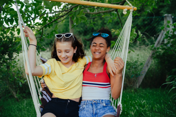 Yong teenage girls friends outdoors in garden, swinging on swing, having fun, talking during warm summer evening.