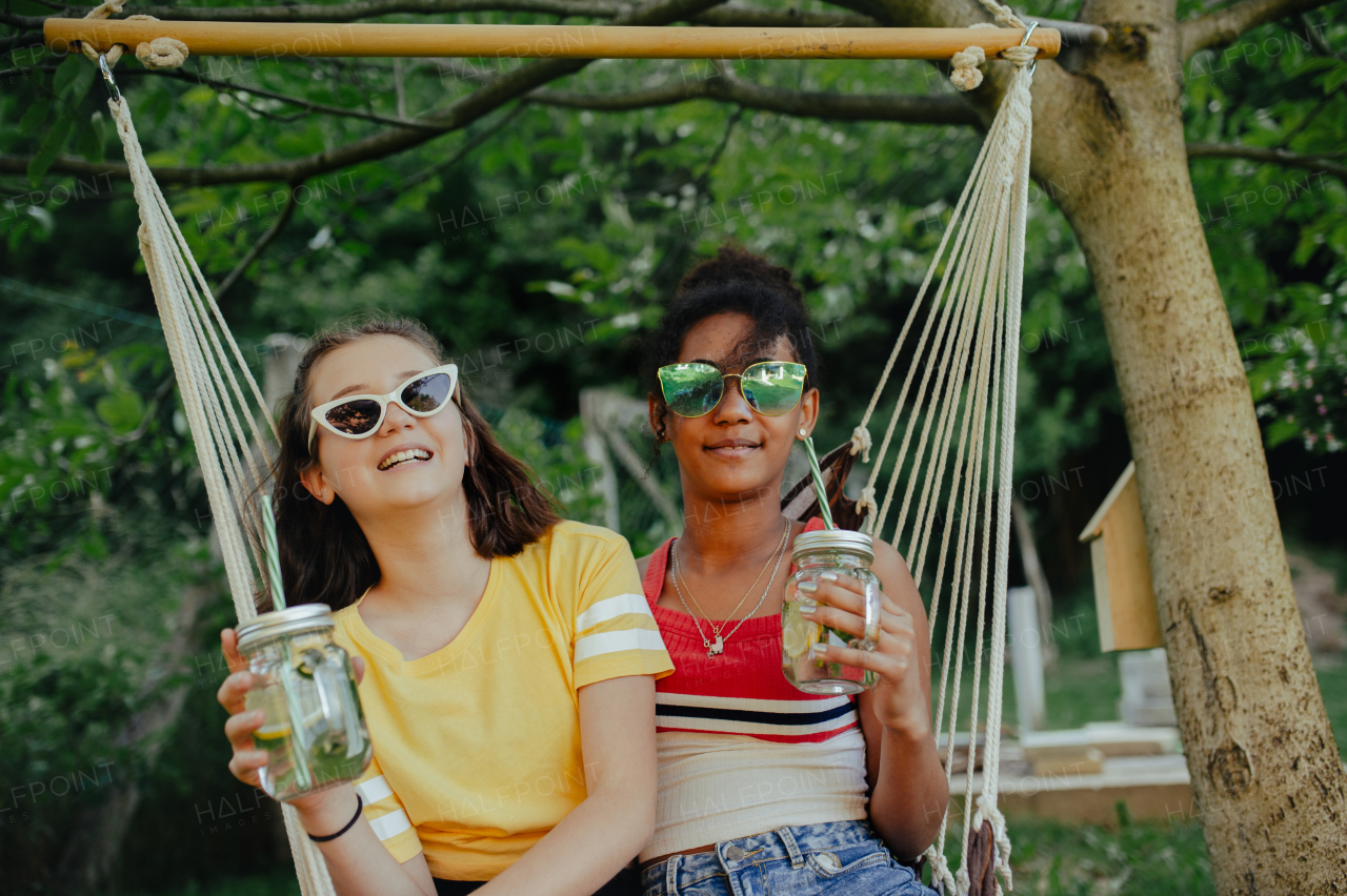 Yong teenage girls friends outdoors in garden, swinging on swing, drinking lemonade from jar glass, during warm summer evening.