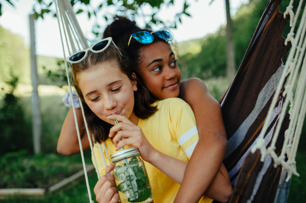 Yong teenage girls friends outdoors in garden, swinging on swing, drinking lemonade from jar glass, during warm summer evening.