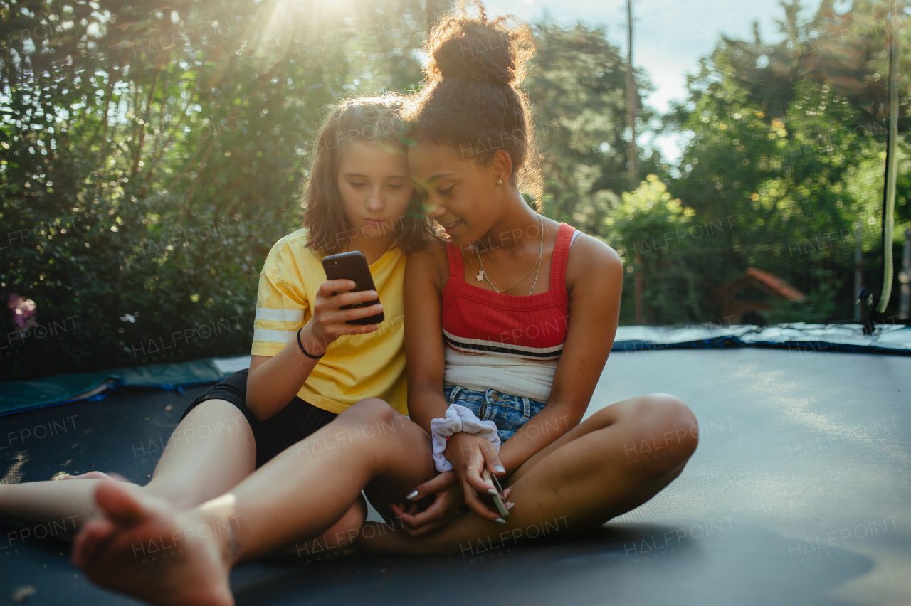 Teenager girls friends spending time outdoors in garden, laughing. Sitting on trampoline, scrolling on smartphone, social media, during warm summer day.