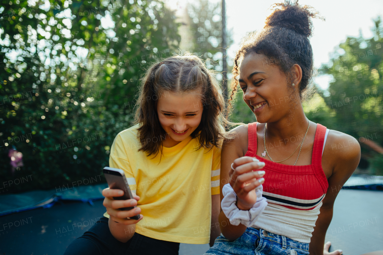 Teenager girls friends spending time outdoors in garden, laughing. Sitting on trampoline, scrolling on smartphone, social media, during warm summer day.