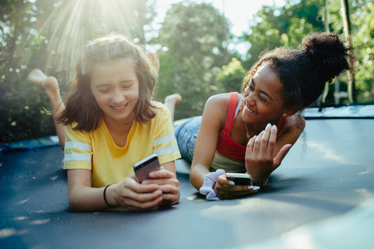 Teenager girls friends spending time outdoors in garden, laughing. Lying on trampoline, scrolling on smartphone, social media, during warm summer day.