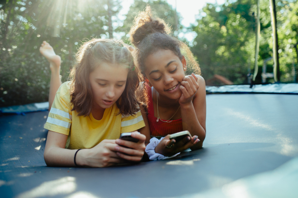 Teenager girls friends spending time outdoors in garden, laughing. Lying on trampoline, scrolling on smartphone, social media, during warm summer day.