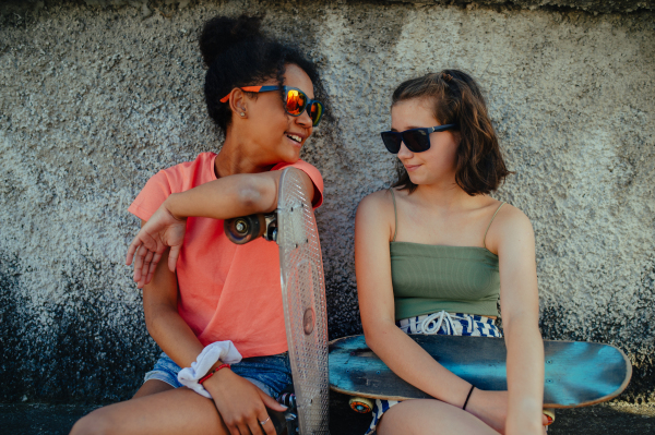 Young teenager girl best friends with skateboards spending time outdoors in city during warm summer holiday day. Sitting by concrete wall.