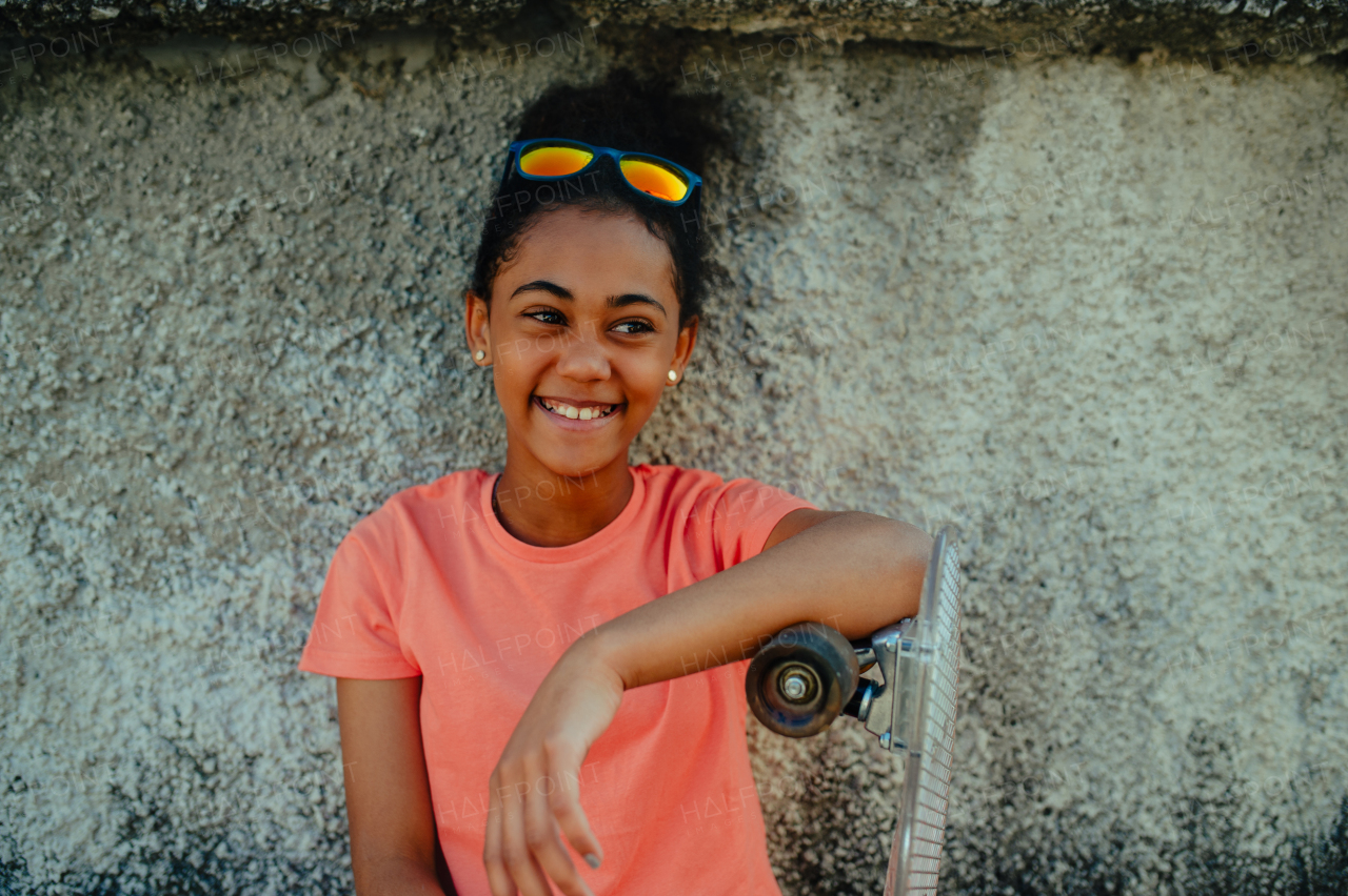 Young teenager girl with skateboards spending time outdoors in city during warm summer holiday day. Sitting by concrete wall.