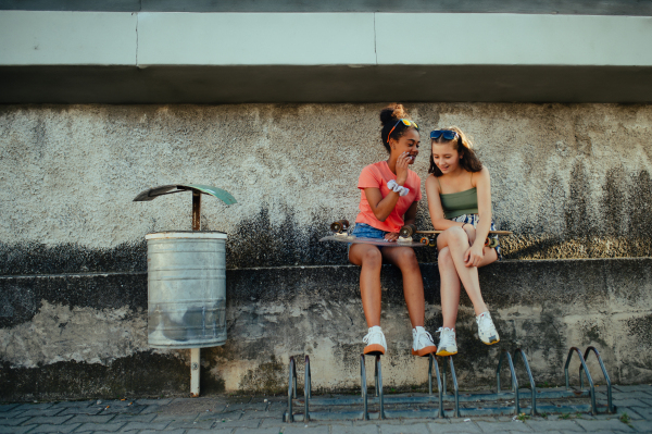 Young Teenager girl best friends with skateboards spending time outdoors in city during warm summer holiday day. Sitting on concrete wall, talking, sharing secret.