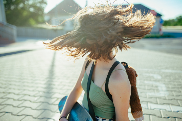 Young teenager girl with skateboards spending time outdoors in city during warm summer holiday day. Sitting on parking lot. Flying hair, spinning head.