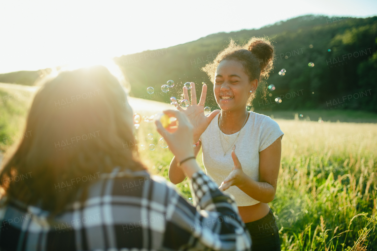 Cheerful young teenager girl best friends spending time in nature, during sunset. Girls blowing bubbles and popping them.