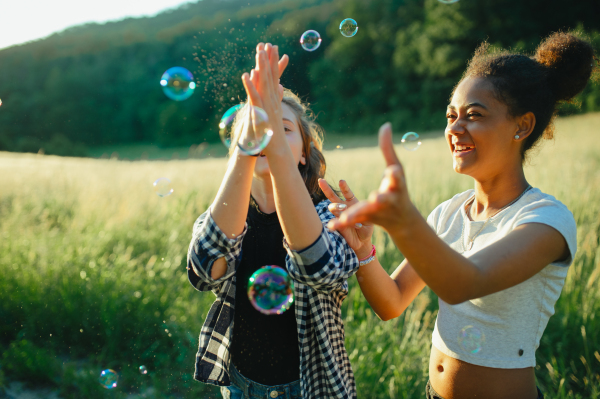 Cheerful young teenager girl best friends spending time in nature, during sunset. Girls blowing bubbles and popping them.