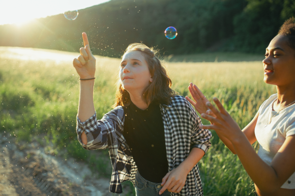 Cheerful young teenager girl best friends spending time in nature, during sunset. Girls blowing bubbles and popping them.