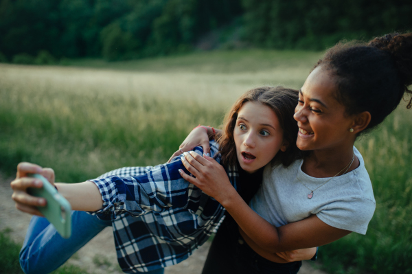 Cheerful young teenager girl best friends spending time in nature, during sunset. Girls on walk, taking selfie with smartphone.