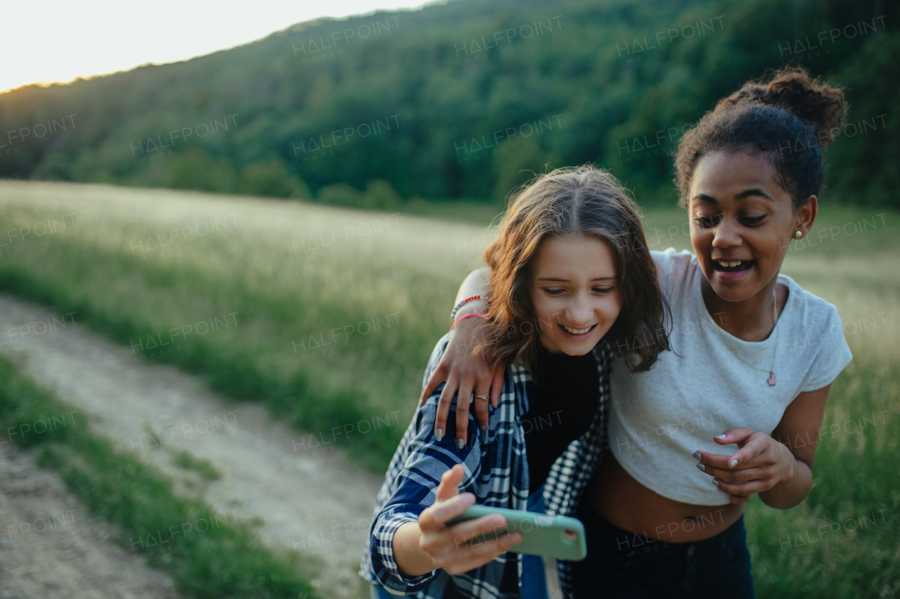 Cheerful young teenager girl best friends spending time in nature, during sunset. Girls on walk, taking selfie with smartphone.