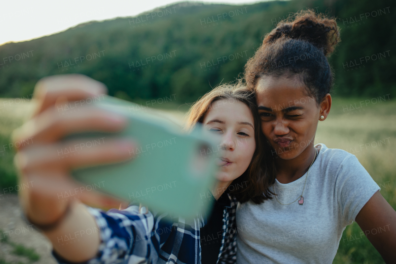 Cheerful young teenager girl best friends spending time in nature, during sunset. Girls on walk, taking selfie with smartphone.