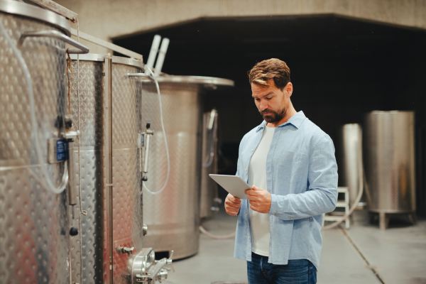 The winemaker standing in the wine cellar, monitoring the fermentation and aging processes in large barrels, holding clipboard.