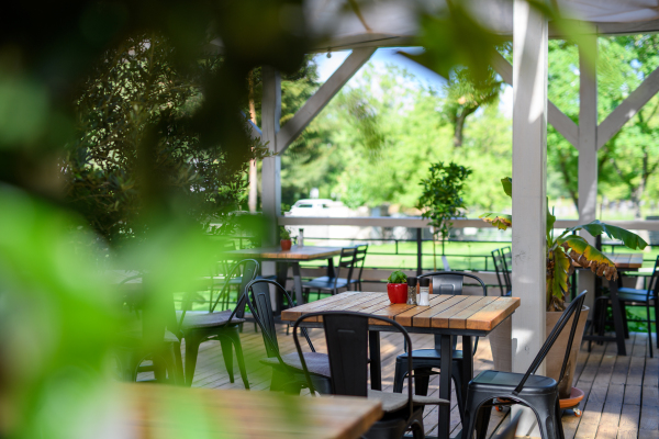 Covered summer terrace of a restaurant with wooden tables, chairs, and flooring. Restaurant patio made of natural materials with lot of greenery.