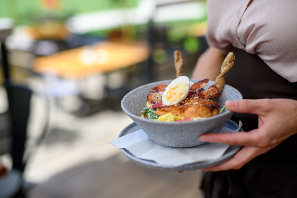 Close up of waitress holding plate with food, bowl with vegetable salad. Server standing on restaurant terrace.