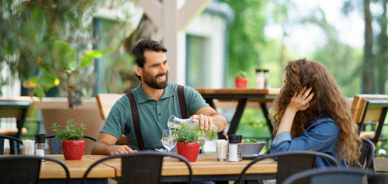 Young couple at date in restaurant, sitting on restaurant terrace. Boyfriend pouring water in girlfriends glass, taking care of her. Lunch or brunch outdoors, outdoor seating for dining. Banner with copy space.