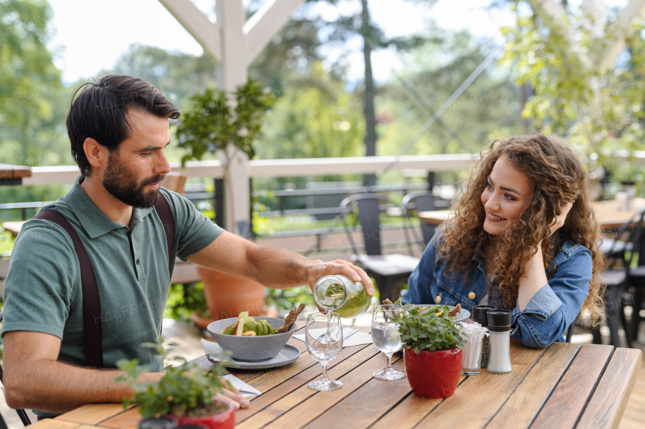 Young couple at date in restaurant, sitting on restaurant terrace. Boyfriend pouring water in girlfriends glass, taking care of her. Lunch or brunch outdoors, outdoor seating for dining.