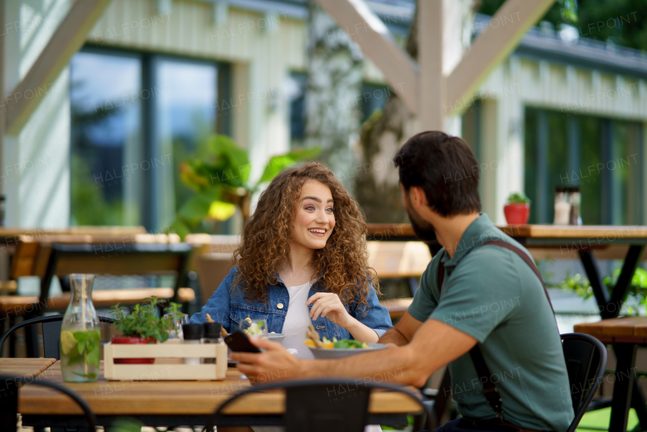 Young couple laughing at date in restaurant, sitting on restaurant terrace. Boyfriend and girlfriend enjoying springtime, having lunch or brunch outdoors, outdoor seating for dining.