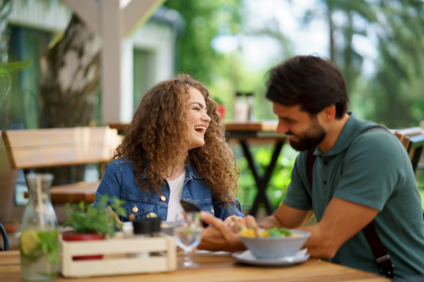 Young couple laughing at date in restaurant, sitting on restaurant terrace. Boyfriend and girlfriend enjoying springtime, having lunch or brunch outdoors, outdoor seating for dining.