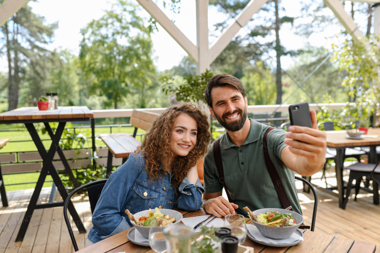 Young couple at date in restaurant, taking selfie, sitting on restaurant terrace. Boyfriend and girlfriend enjoying springtime, having lunch or brunch outdoors, outdoor seating for dining.