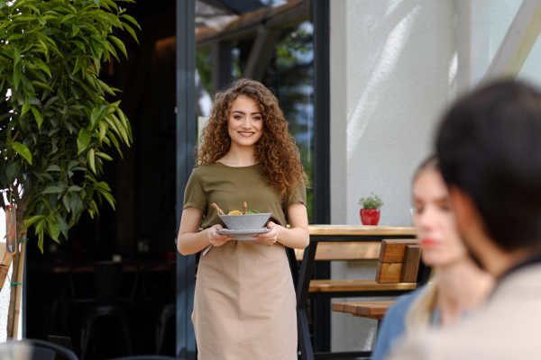 Portrait of a beautiful waitress holding plate with food, bowl with salad. Female server in apron bringing food to customers on restaurant terrace.