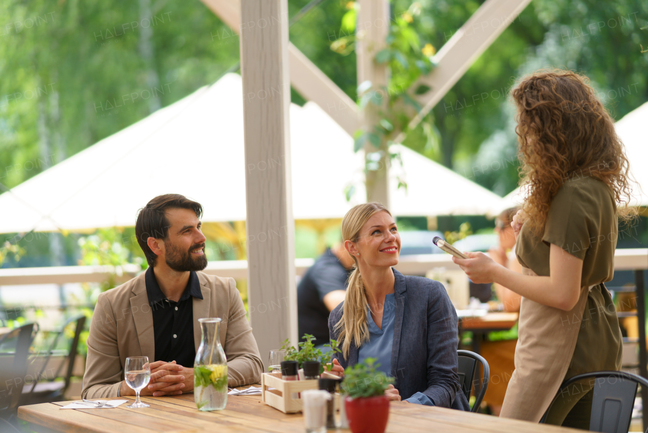 Couple sitting outdoors on terrace restaurant, ordering food from female waiter. Business lunch for two managers, discussing new business project.