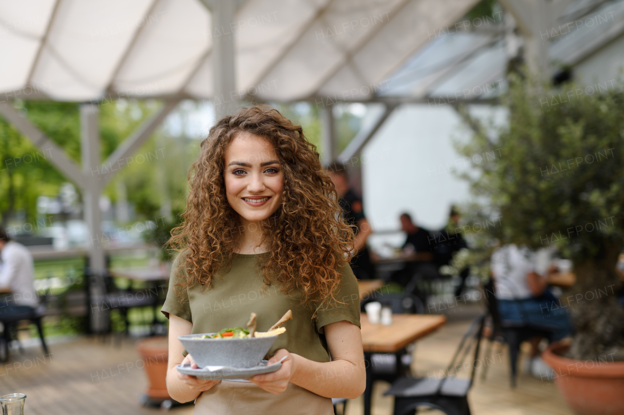 Portrait of a beautiful waitress holding plate with food, bowl with salad.Female server standing on the restaurant terrace in an apron, looking at camera, smiling.