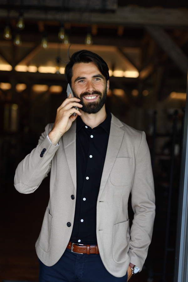 Portrait of mature businessman in suit jacket standing indoors in the modern office, making phone call.