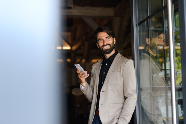 Portrait of mature businessman in suit jacket standing indoors in modern office, holding smartphone in hand.