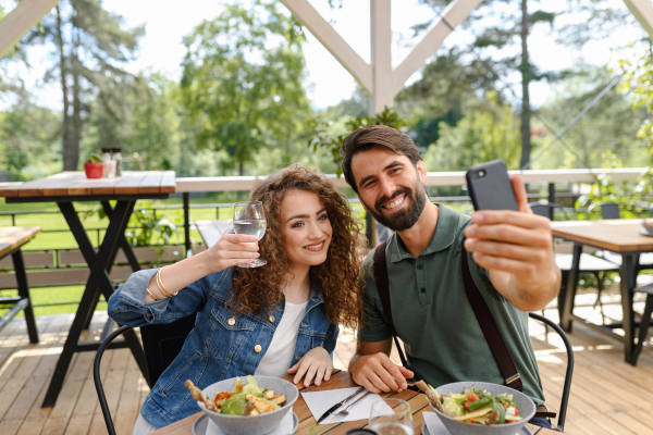 Young couple at date in restaurant, taking selfie, sitting on restaurant terrace. Boyfriend and girlfriend enjoying springtime, having lunch or brunch outdoors, outdoor seating for dining.