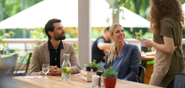 Couple sitting outdoors on terrace restaurant, ordering food from female waiter. Business lunch for two managers, discussing new business project.