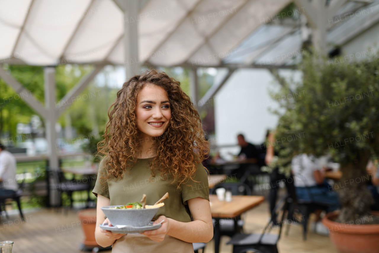 Portrait of a beautiful waitress holding plate with food, bowl with salad.Female server standing on the restaurant terrace in an apron.