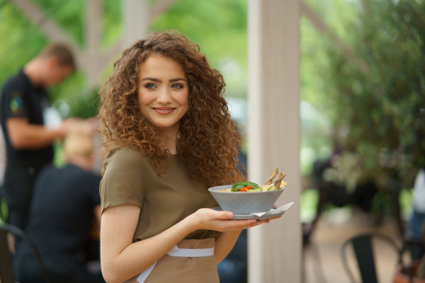 Portrait of a beautiful waitress holding plate with food, bowl with salad.Female server standing on the restaurant terrace in an apron.