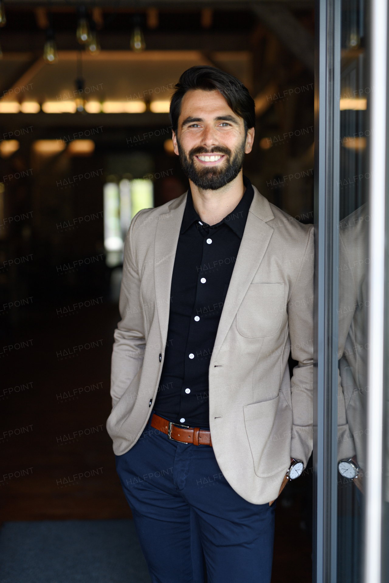 Portrait of mature businessman in suit jacket standing indoors in modern office, looking at camera, smiling.