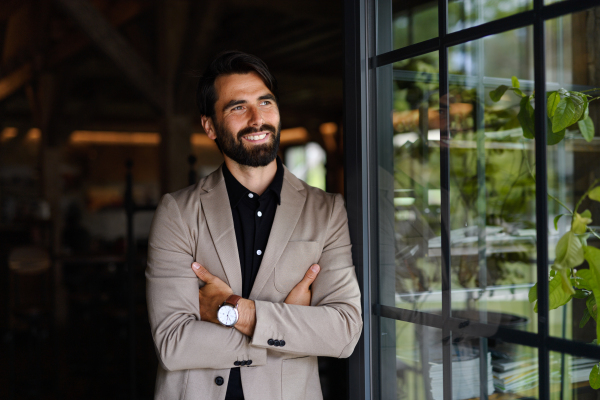 Portrait of mature businessman in suit jacket standing indoors in modern office, looking out.