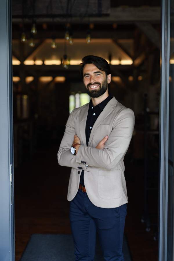 Portrait of mature businessman in suit jacket standing indoors in modern office, looking at camera, smiling.