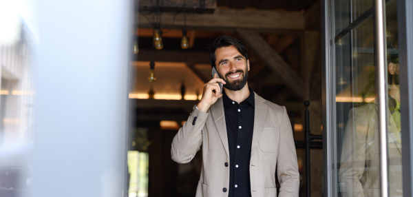 Portrait of mature businessman in suit jacket standing indoors in the modern office, making phone call. Banner with copyspace.