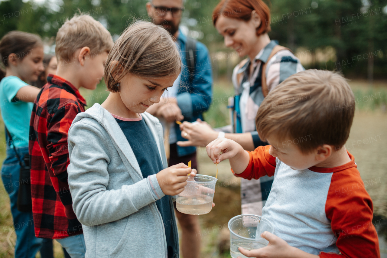 Young students analyzing water quality, ph level with indicator strips during biology field teaching class. Female teacher during outdoor active education about ecosystem, nature.