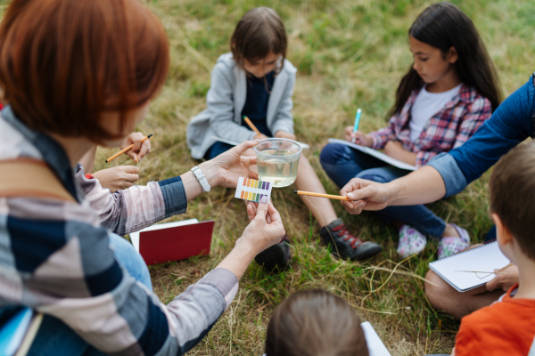 Young students analyzing water quality, ph level with indicator strips during biology field teaching class. Female teacher during outdoor active education about ecosystem, nature.