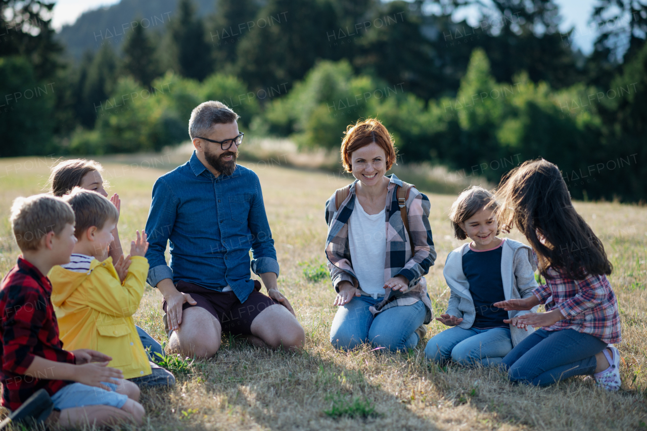 Children and teachers sitting on grass on meadow playing clapping game. Dedicated teachers during outdoor active education teaching about ecosystem, ecology and nature.