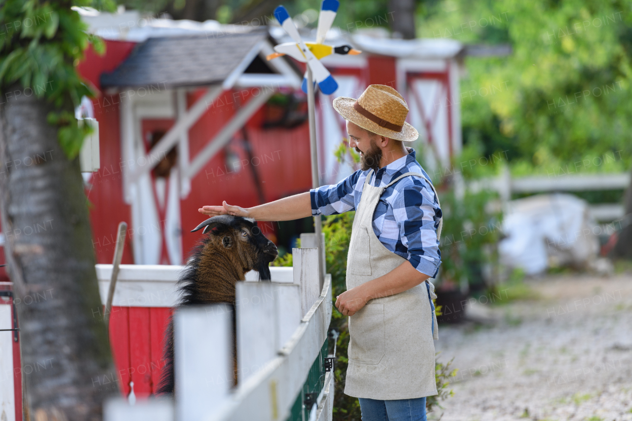 Portrait of handsome farmer petting brown goat at his farm. Man with hat working on family ranch.