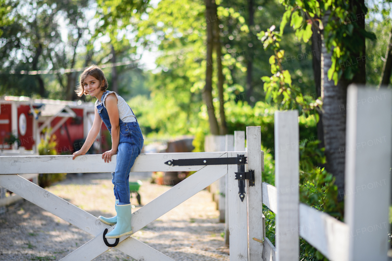 Beautiful young girl in rubber boots and overalls sitting on white wooden fence, helping on family farm during summer.