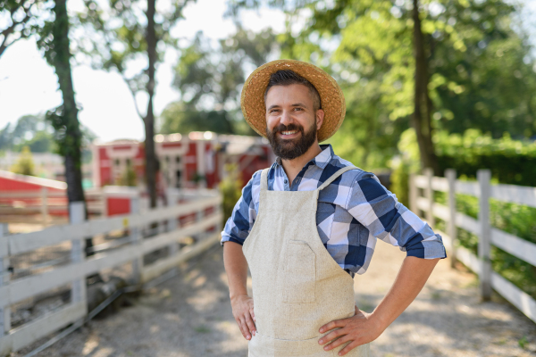 Portrait of handsome farmer standing by wooden fence, looking at his farm. Man with hat working on family ranch.