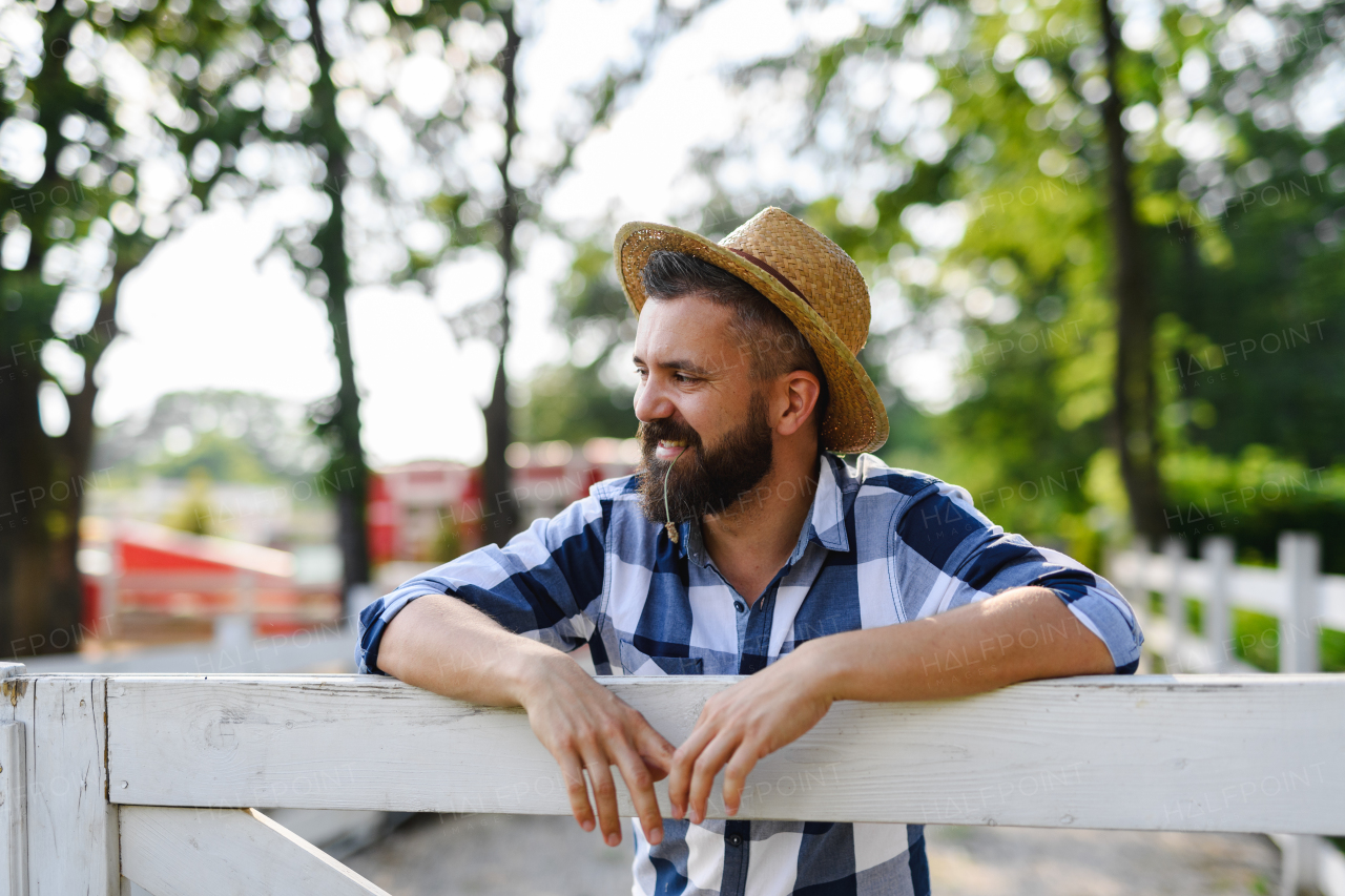 Portrait of handsome farmer leaning agaist wooden fence, looking at his farm. Man with hat working on family ranch.